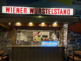 Ein Würstelstand in Wien bei Nacht. Die Aufschrift zeigt "Wiener Würstelstand". Hinter der Theke arbeiten ein Mann und eine Frau