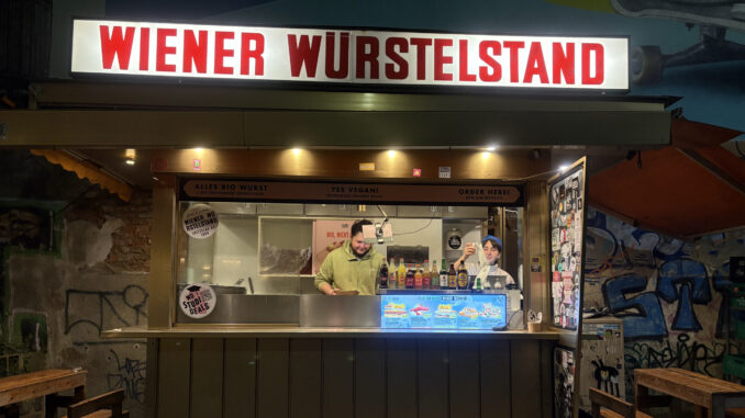 IMG_5508 Ein Würstelstand in Wien bei Nacht. Die Aufschrift zeigt "Wiener Würstelstand". Hinter der Theke arbeiten ein Mann und eine Frau